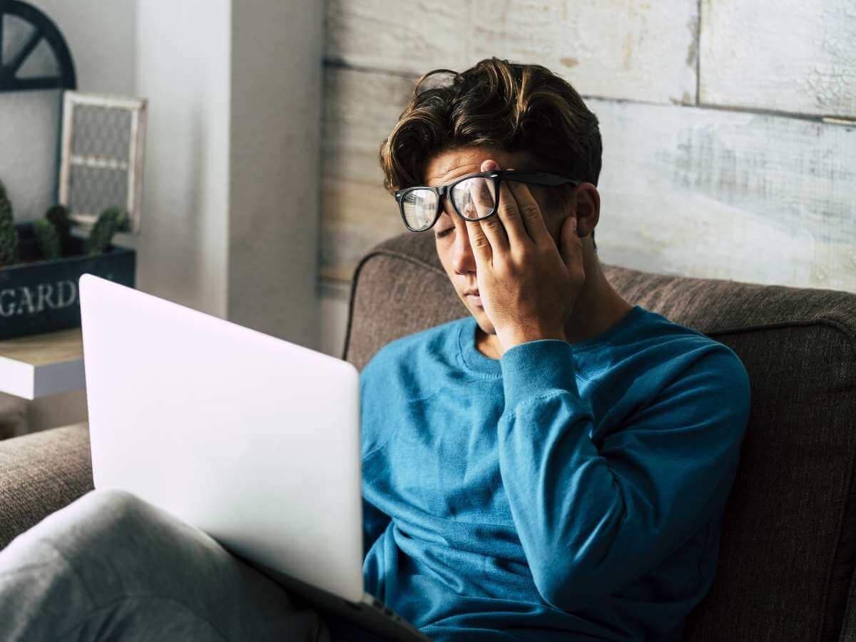 A stressed man wearing a blue shirt sitting on the sofa using a laptop computer.