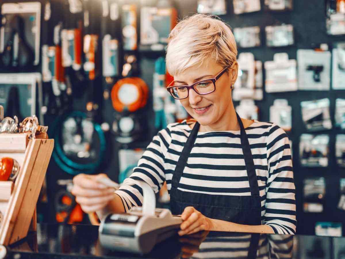 Smiling female salesperson with short blonde hair and eyeglasses using cash register while standing in bicycle store.