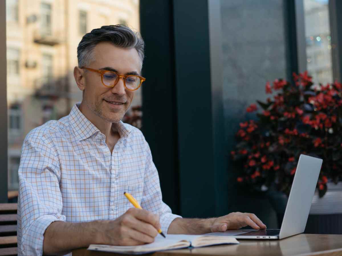 Portrait of a middle aged man in orange glasses sitting outside with a laptop and taking notes