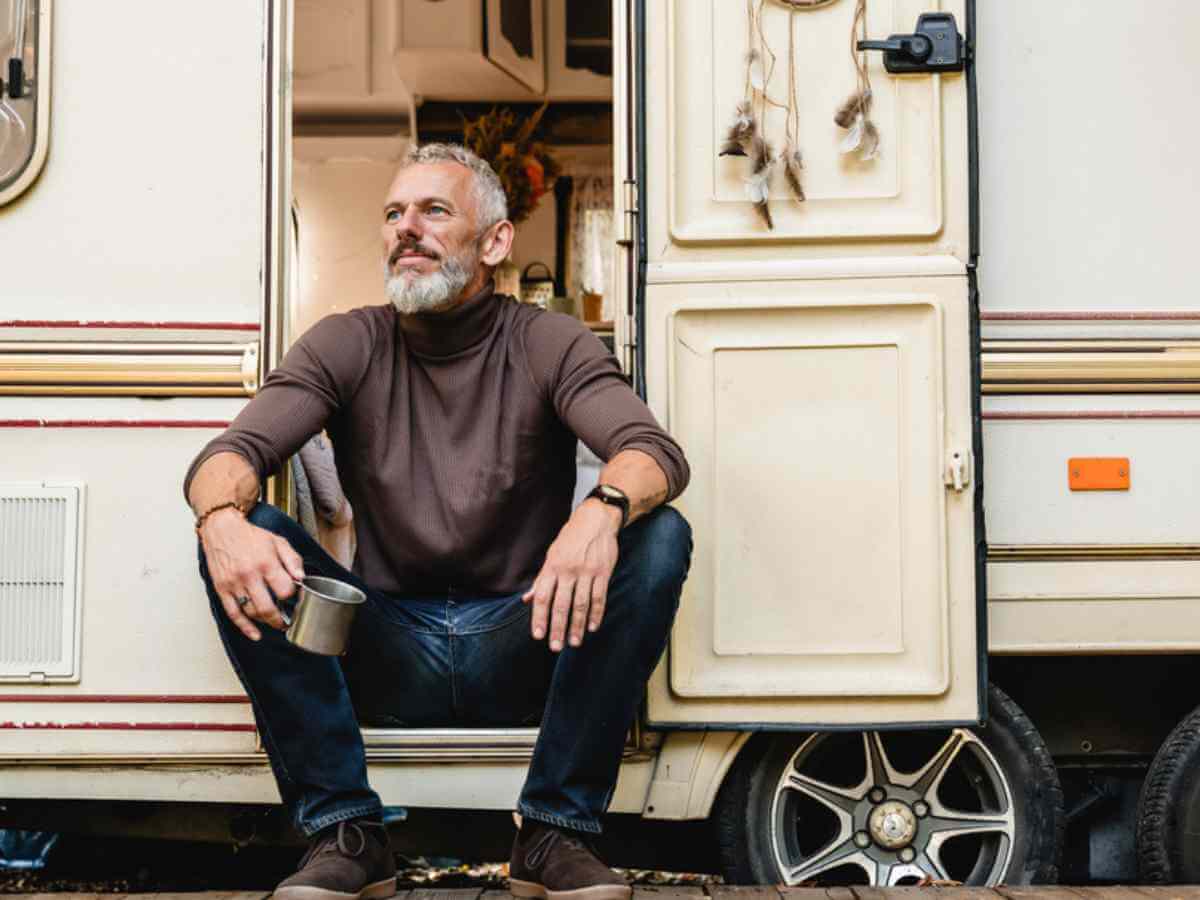 Portrait of handsome senior man with a beard sitting on the doorstep of motor home holding an enamel camping mug