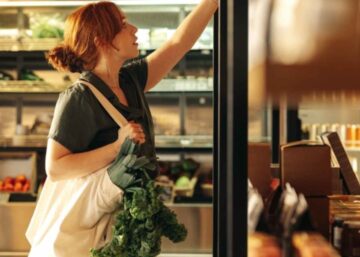 Female customer in a grocery store choosing food products from a shelf while carrying a bag containing vegetables.