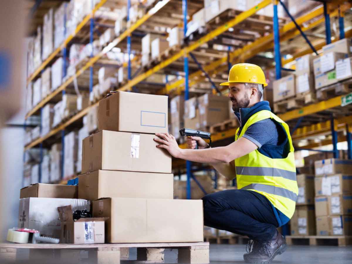 A male warehouse worker in a yellow vest and hard hat kneels in front of a pile of cardboard boxes with a barcode scanner.