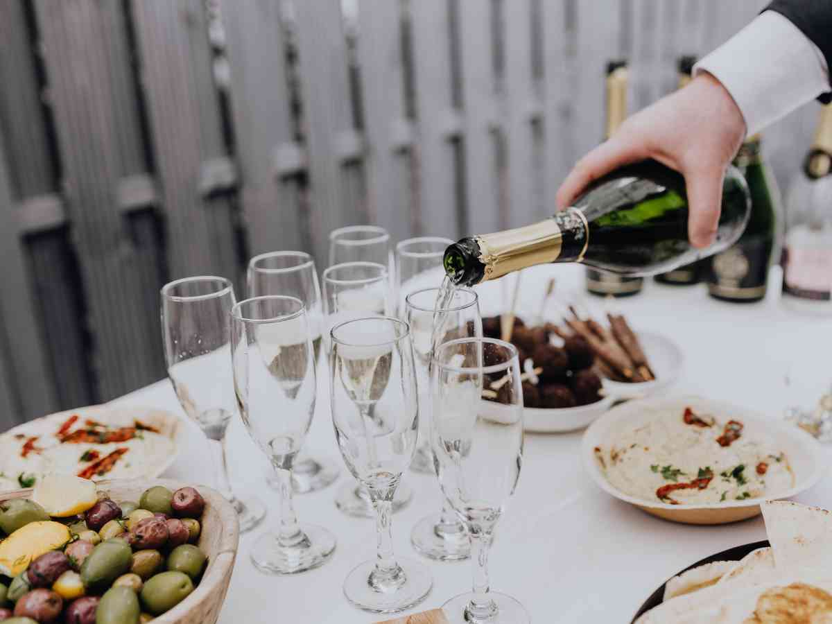 Close up of a waiter pouring champagne into glasses set up on a buffet table outside with plates of olives and canapes.