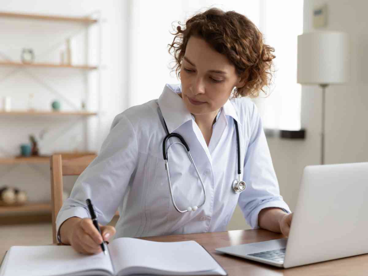 Serious female doctor wearing a white coat and stethoscope using laptop and writing notes while sitting at desk